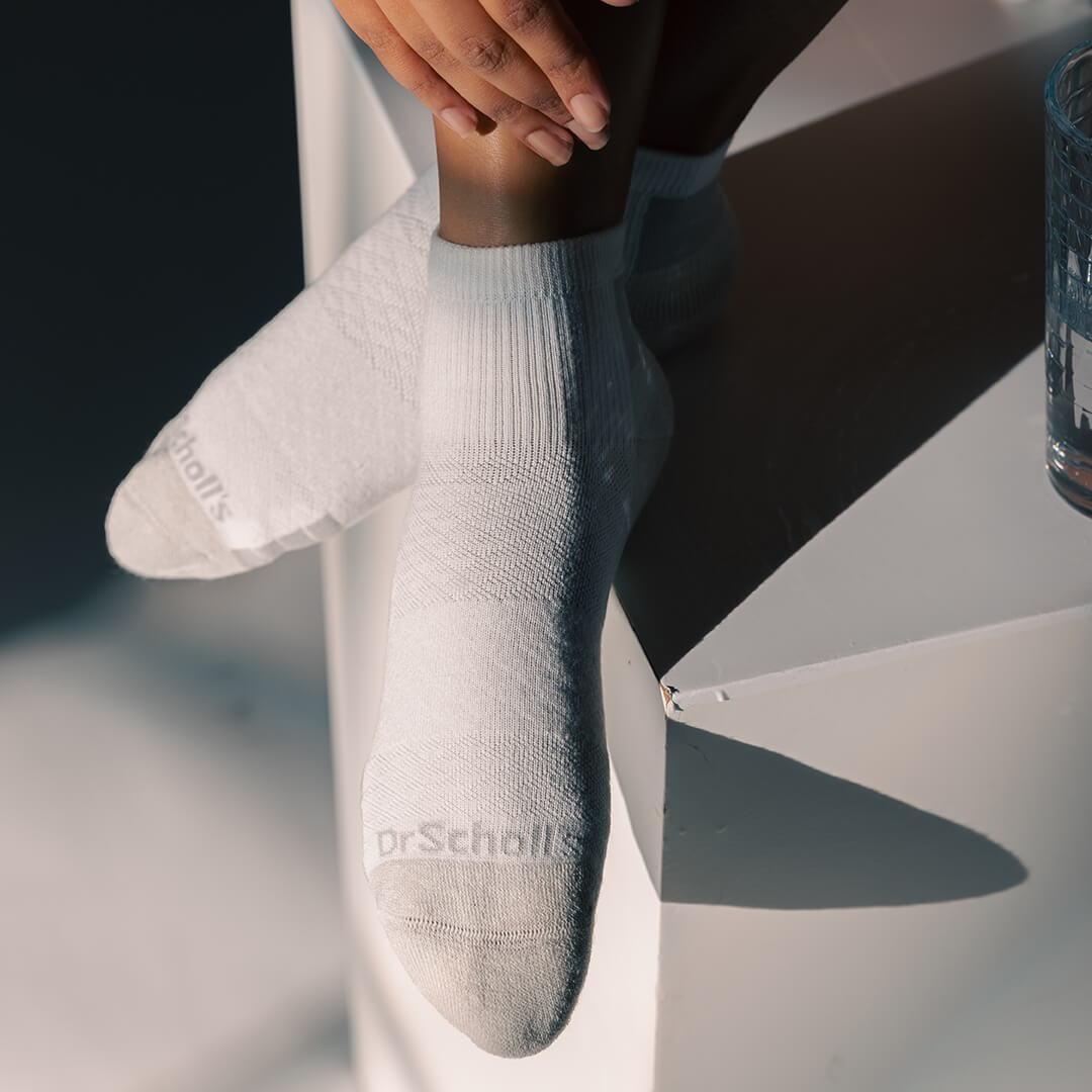 A close-up of a person's foot and ankle wearing a light gray ribbed ankle sock with 'DrScholl's' printed on the toe, resting on a white geometric surface.