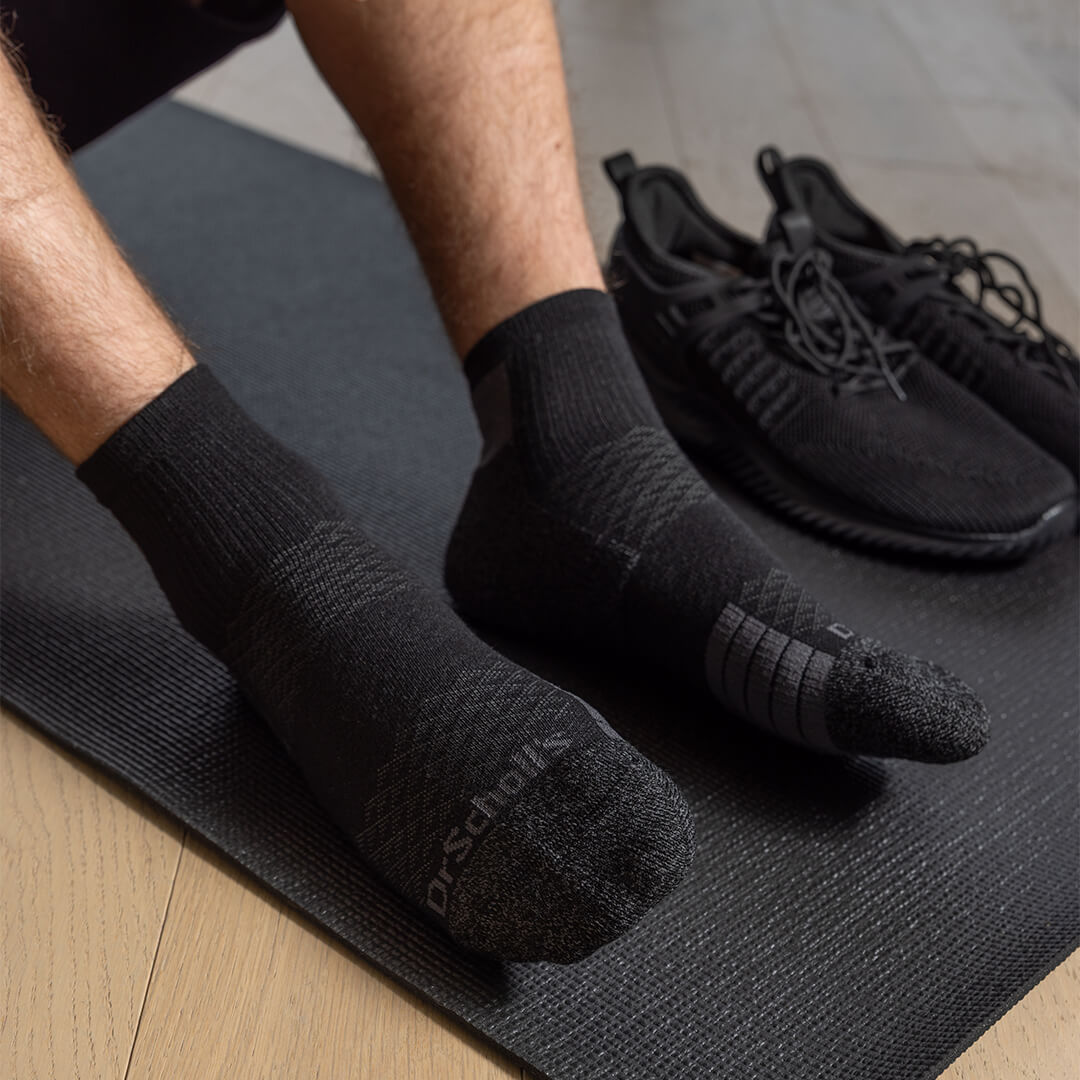 A person wearing black Dr. Scholl's athletic socks sits on a black yoga mat with black sneakers in the background.
