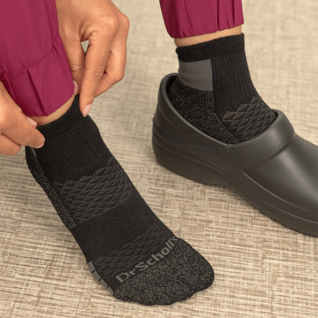 A close-up of a person wearing black Dr. Scholl's socks with textured knit patterns, shown being put on and worn with black clogs on a textured beige floor.