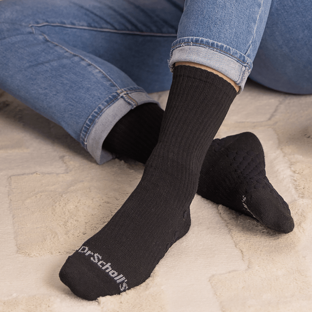 A person wearing black crew socks with the brand name 'Dr. Scholl' visible on the sole of one sock, seated on a light beige textured rug with blue jeans visible.