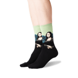 A pair of crew socks worn on a person's feet, featuring a black lower half and a light green upper half with a printed parody of the Mona Lisa wearing sunglasses.