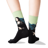 A pair of crew socks worn on a person's legs, featuring a black lower half with a printed Mona Lisa design and a light green upper half with blue mountain graphics. thumbnail