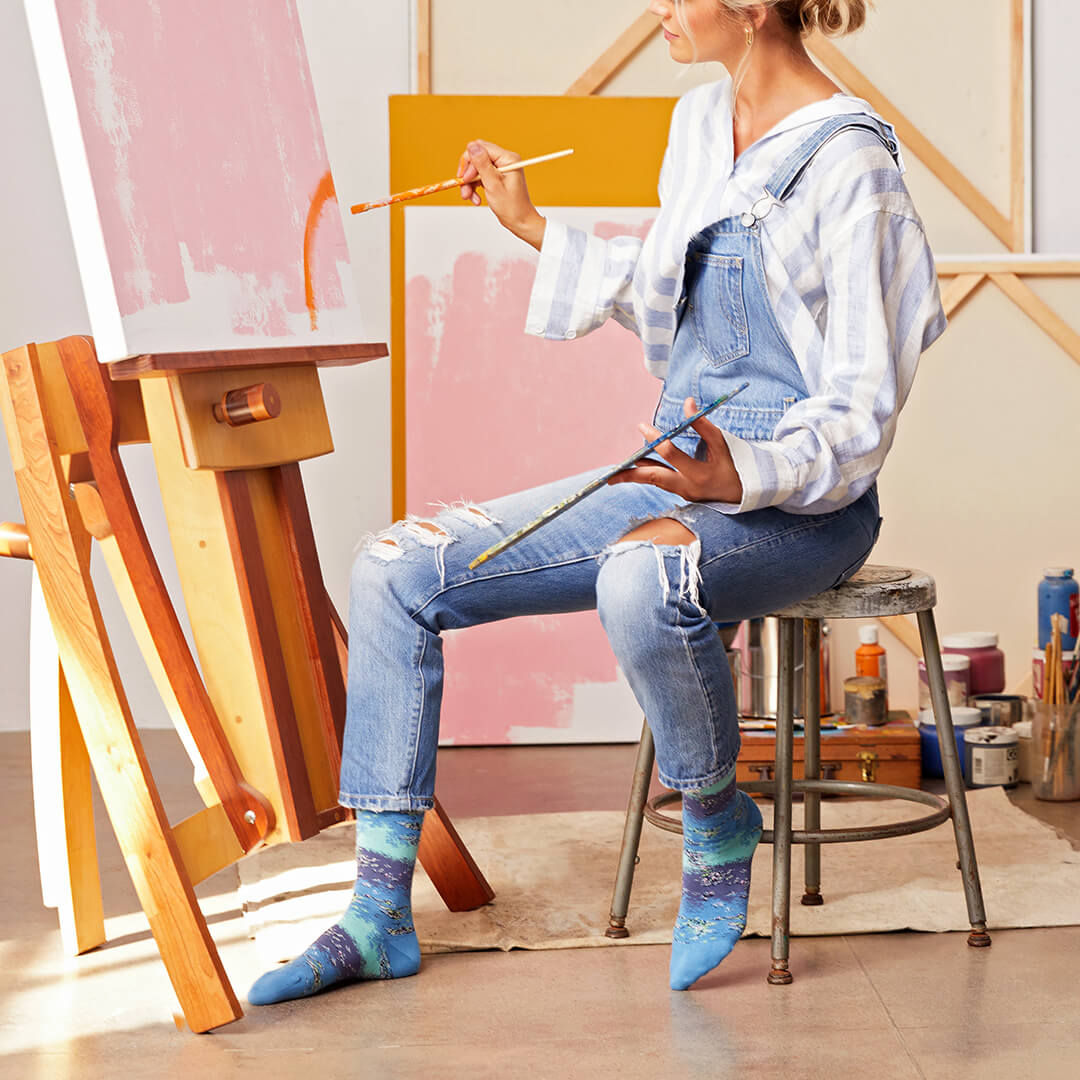 A woman sitting on a metal stool in an art studio, wearing blue denim overalls and striped blue-and-white long-sleeve shirt, holding paintbrushes. She is positioned next to a wooden easel with pink and orange paint on the canvas, with other art supplies on a stool behind her.