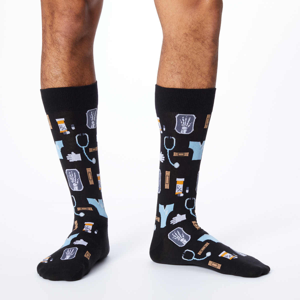 A pair of black crew socks with a medical-themed print of stethoscopes, X-rays, pills, and hands, shown on a model's legs against a white studio background.