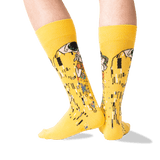 A pair of bright yellow crew socks with a Gustav Klimt-style art print featuring black, white, green, and orange details, shown on a person's legs against a clean white background. thumbnail