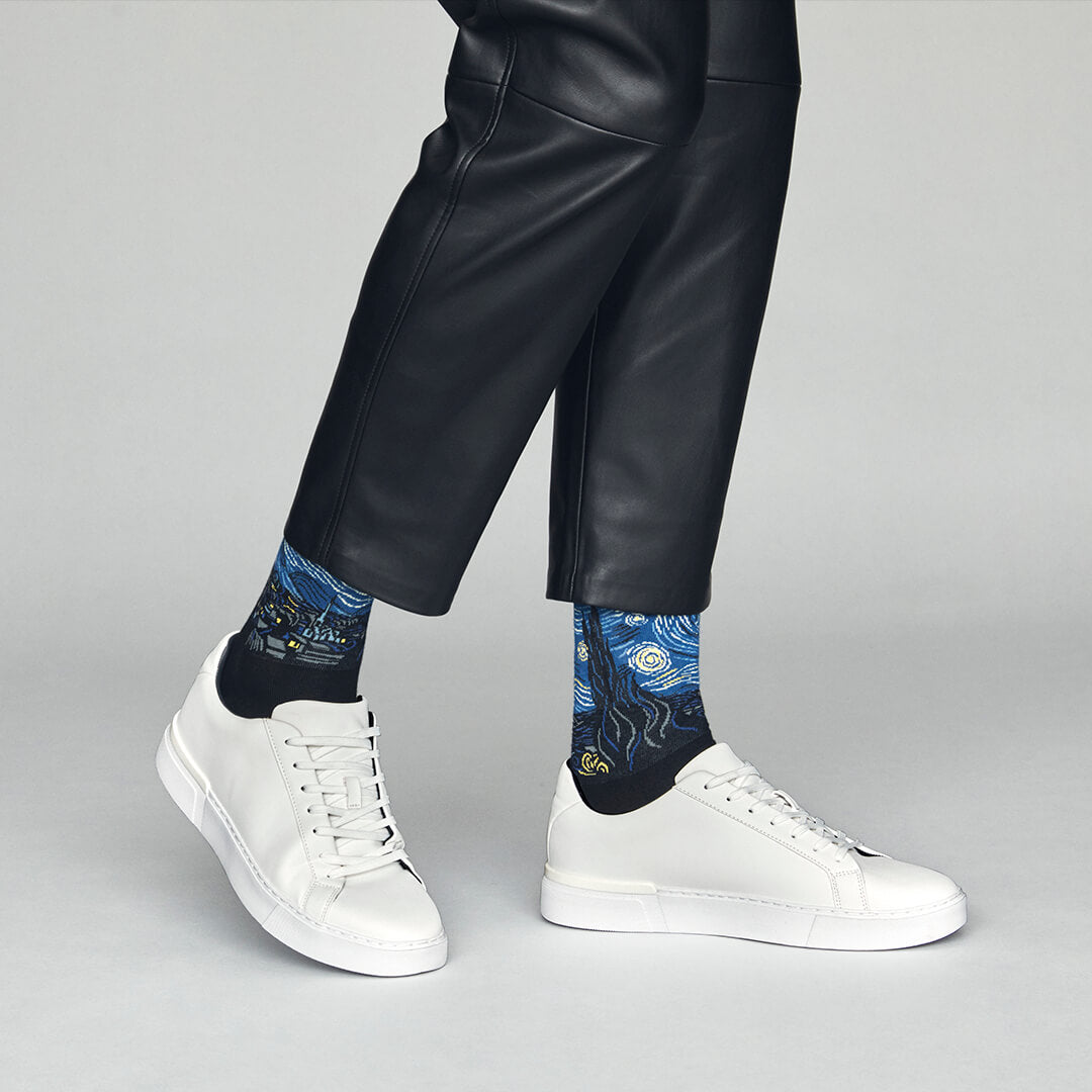 Person wearing black leather pants and white sneakers with blue patterned socks on a gray background