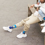 A man sitting on a wooden bench outdoors on a gray, hexagonal-paved surface, wearing beige cargo pants, white sneakers, and bright blue crew socks with Van Gogh's "Self Portrait" knit into the design.