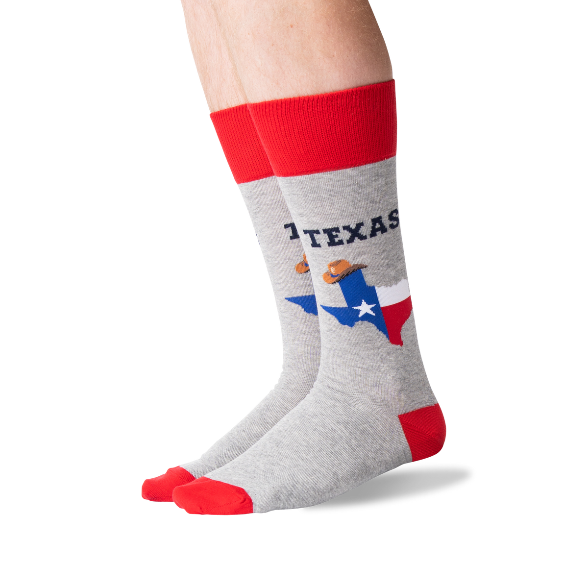 A pair of crew socks worn on a person's legs, featuring a heather gray body with a red cuff, red toe, and red heel. The socks display 'TEXAS' in navy blue text above a graphic of the state of Texas filled with the Texas flag and a cowboy wearing a sombrero.