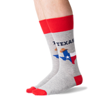 A pair of crew socks worn on a person's legs, featuring a heather gray body with a red cuff, red toe, and red heel. The socks display 'TEXAS' in navy blue text above a graphic of the state of Texas filled with the Texas flag and a cowboy wearing a sombrero. thumbnail