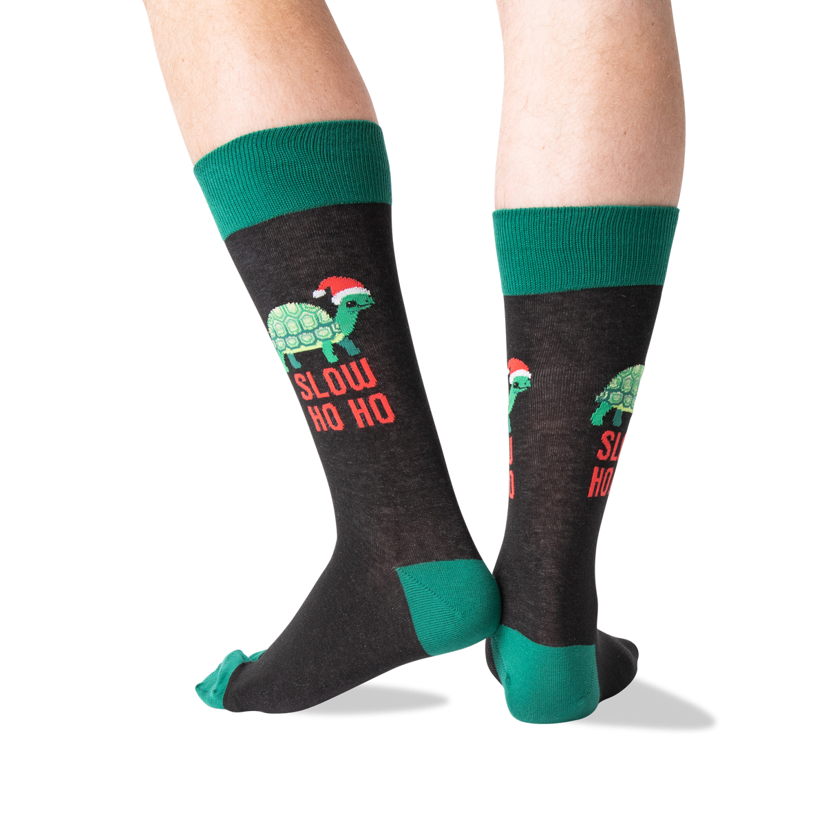 A pair of crew socks worn on a person's legs, featuring a dark charcoal gray body with green cuffs, heels, and toes, and a graphic of a green turtle wearing a red Santa hat with the text 'SLOW HO HO' in red.