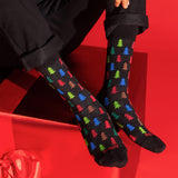 A person wearing black crew socks with a colorful Christmas tree pattern, posed against a bright red background with a red gift box. thumbnail