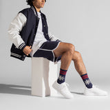 A model sits on a white block wearing a navy and white varsity jacket, matching shorts, white sneakers, and navy crew socks with red stripes and white snowflake patterns. thumbnail