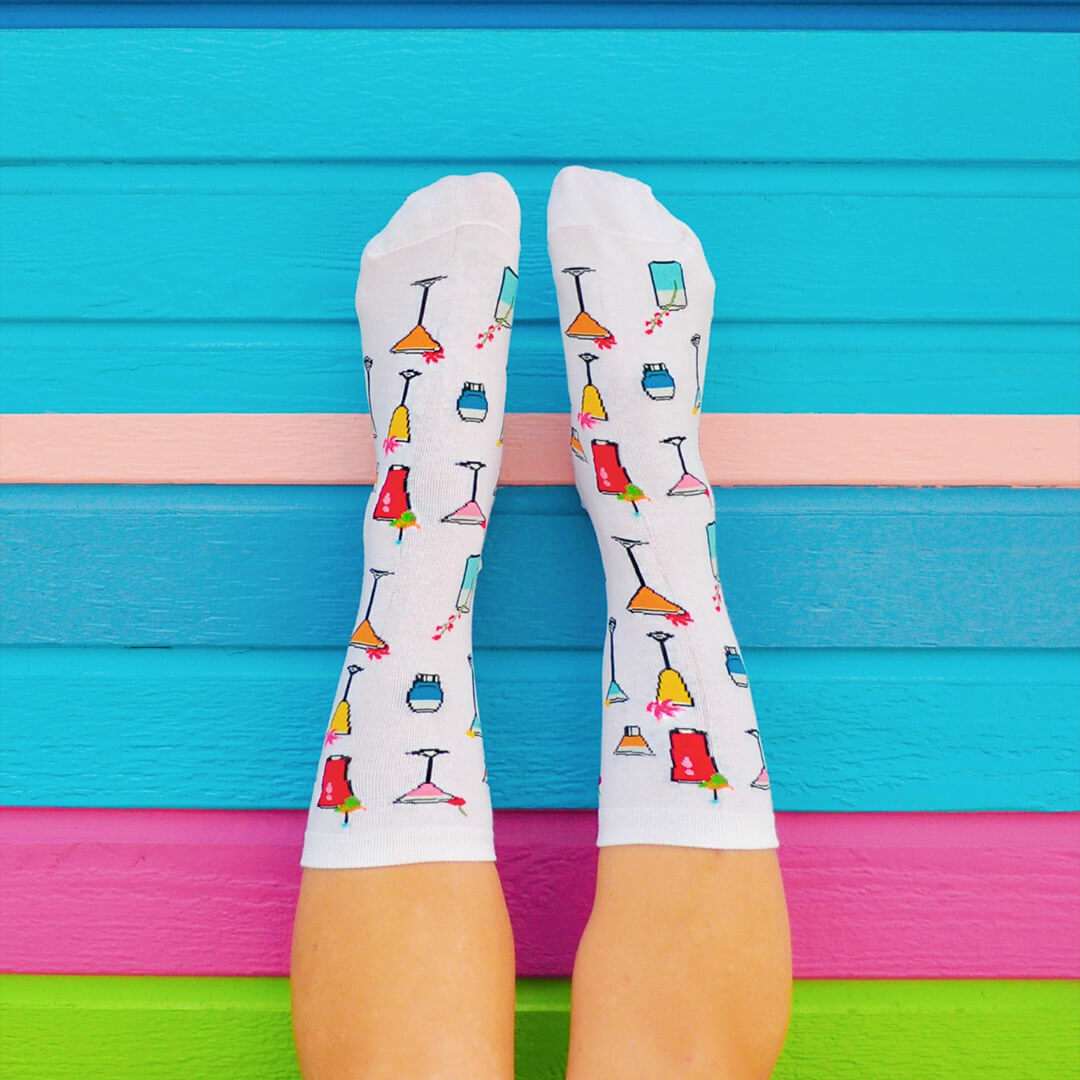 A pair of white crew socks with a colorful cocktail glass pattern, worn on feet against a bright blue wooden background with pink and green stripes.