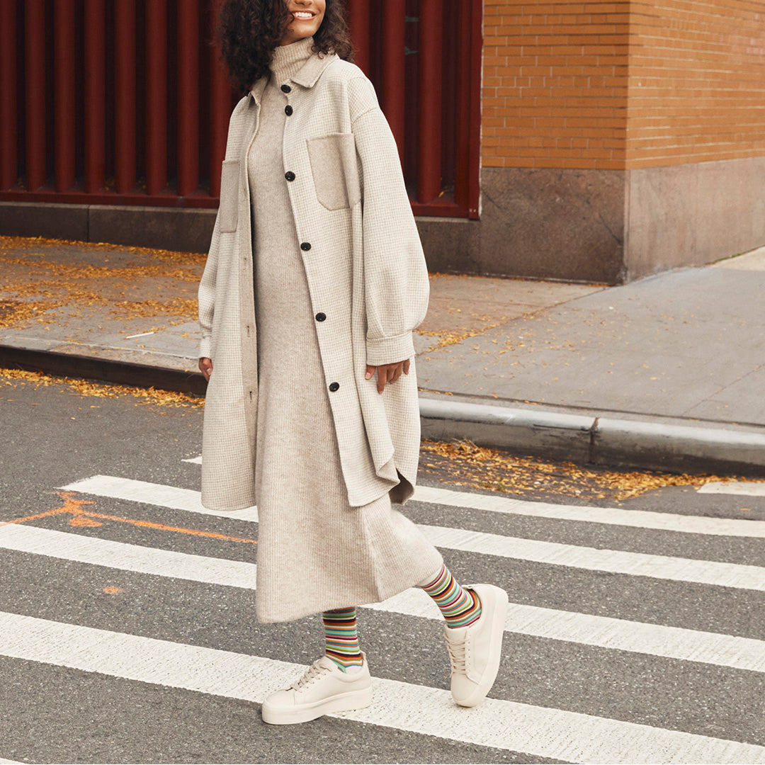 A woman walking across a city crosswalk wearing an oversized light beige button-front coat over a matching long-sleeve knit dress, with striped socks and white sneakers.