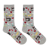 A pair of heather gray crew socks with an all-over sewing-themed pattern featuring black sewing machines, red hearts, yellow spools, scissors, and other colorful thread icons, shown against a plain white background.