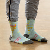 A person wearing light blue crew socks with a yellow menorah, pink candles, and pink text, paired with gray sweatpants, standing on a wooden floor.