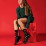 A woman sits on a large red gift box against a solid red studio background, wearing a dark green cable-knit sweater dress, black leather shoes, and black crew socks with a colorful Christmas tree pattern. thumbnail