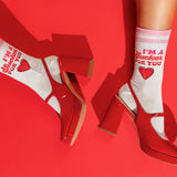 White crew socks with red text reading 'I'M A Sucker FOR YOU' and a red heart, worn with glossy red block-heel shoes against a bright red background.