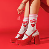 A person seated against a bright red studio background wearing white socks with pink stripes and red text that reads “I’m a Sucker for You,” paired with glossy red Mary Jane-style heels with block heels and ankle straps. thumbnail