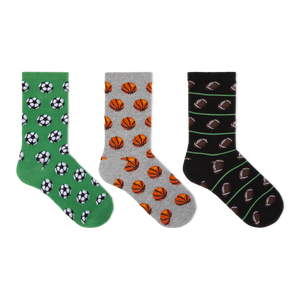Three pairs of crew socks displayed side by side on a plain white background. The left pair is green with black and white soccer ball patterns, the middle pair is gray with orange basketball patterns, and the right pair is black with brown football patterns and thin green stripes.