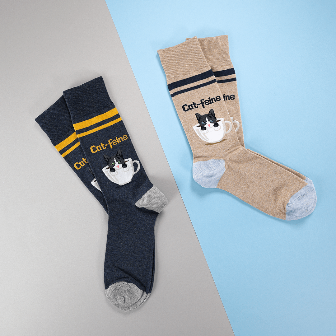 Two pairs of crew socks displayed on a split gray and light blue background. One pair is navy blue with yellow stripes and a cat-in-a-teacup graphic, and the other pair is heather beige with dark navy stripes and a matching cat-in-a-teacup graphic.