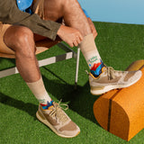 A person seated on a chair outdoors on artificial grass, wearing beige hiking socks with colorful mountain graphics and text, paired with tan and gray sneakers. thumbnail