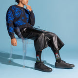 A model sits on a clear acrylic chair wearing a black and blue patterned sweater, black leather-like cropped pants, black sneakers, and black crew socks with colorful graphic prints. The background is a solid light blue studio backdrop. thumbnail