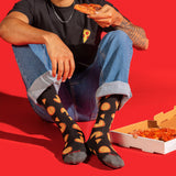 A person sitting on a bright red floor wearing black socks with a repeating pizza slice pattern, holding a slice of pizza. A pizza box is open on the floor beside them.