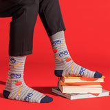 Gray striped socks with a school-themed pattern of blue glasses, yellow pencils, and red apples, worn with black pants and styled on a stack of books against a bright red background. thumbnail
