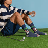 A man sits on artificial grass wearing navy and cream striped polo shorts and dark blue crew socks with a golf-themed graphic. He holds a glass, with a golf club and golf ball nearby against a solid light blue background.