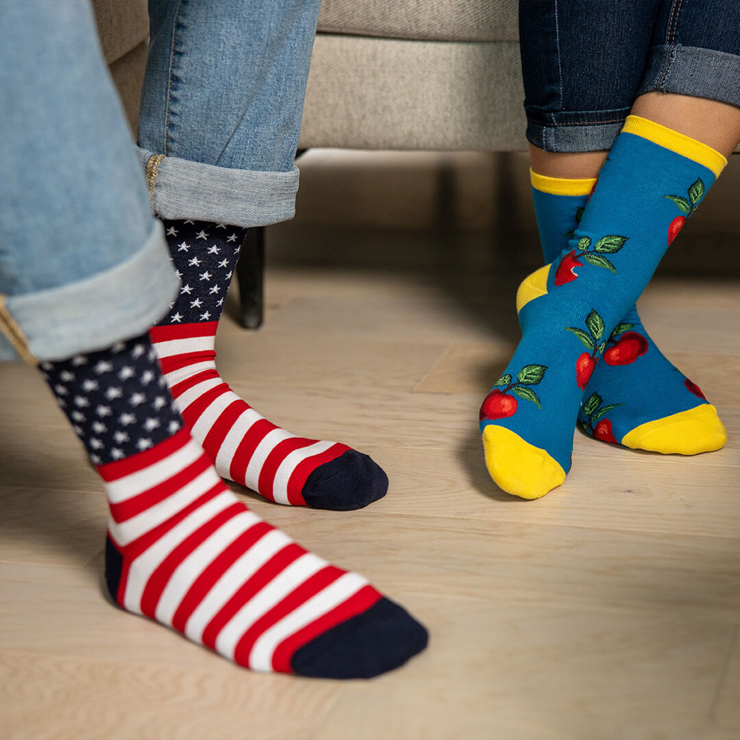 Two people wearing colorful novelty crew socks, one with red and white stripes and a navy star pattern, and the other with a blue cherry print and yellow toes, standing on a light wood floor.