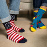 Two people wearing colorful novelty crew socks, one with red and white stripes and a navy star pattern, and the other with a blue cherry print and yellow toes, standing on a light wood floor. thumbnail