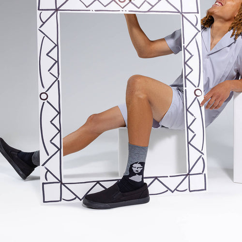 A person in light gray pajamas sits behind a large decorative white frame, wearing black slip-on shoes and a gray crew sock with a black-and-white face graphic. The photo is taken in a clean white studio setting.