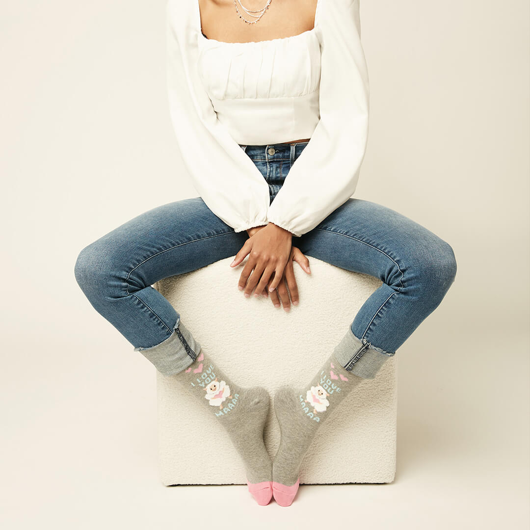 A model sits on a white textured cube sheep gray crew socks with a cute bunny graphic and pink toes, paired with blue jeans and a white long-sleeve top against a light beige background.