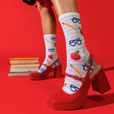 A person wearing white striped socks with red apples, blue glasses, and yellow pencils, paired with red Mary Jane high heels, posed against a solid red background with a stack of books. thumbnail