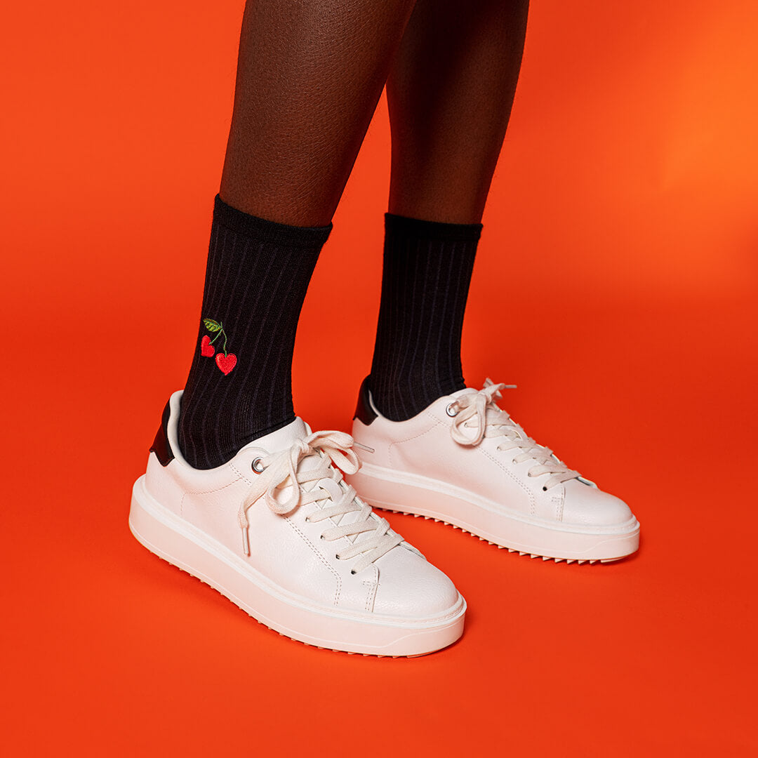 White sneakers worn with black socks featuring a cherry design on an orange background