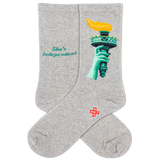 A pair of heather gray crew socks featuring a graphic of the Statue of Liberty's hand holding a yellow torch, with the text 'She's Independent' printed in teal on the other sock. A small red and white 'HOTSOX' logo is visible near the ankle.