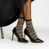 A person seated in a black chair wearing black pointed-toe high heels and metallic-striped crew socks with black ruffled tops. The socks have horizontal stripes in black, mustard yellow, gray, and white. thumbnail