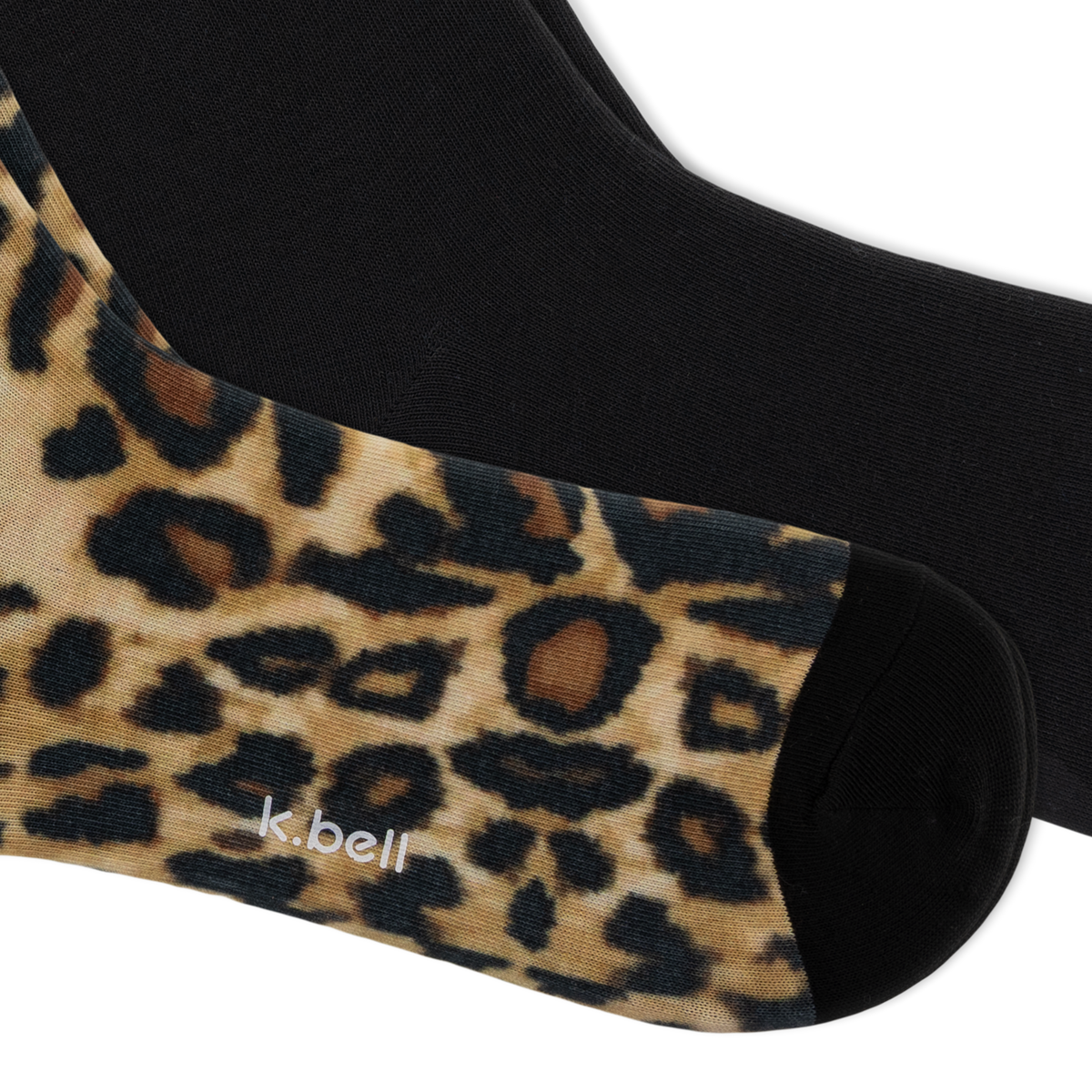 A close-up of a pair of crew socks, with one sock showing a leopard print pattern in beige, brown, and black, and the other sock showing a solid black knit. The brand name 'k.bell' is on the bottom of the foot.