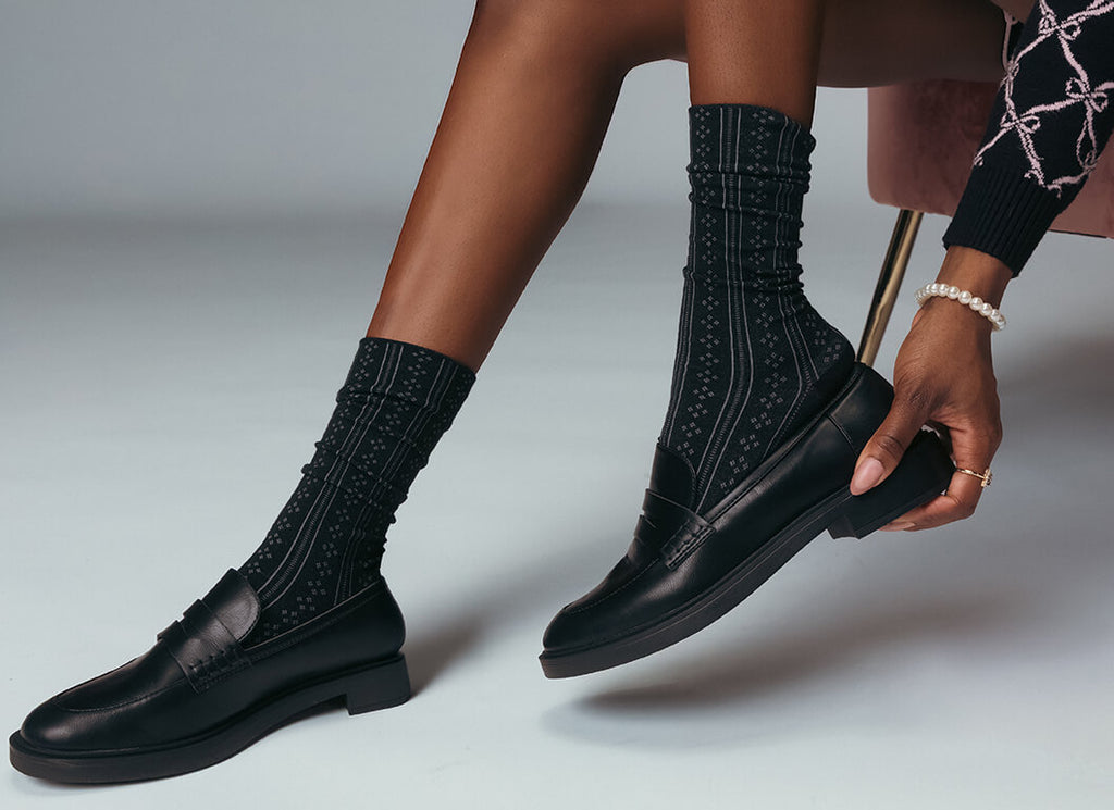 Black dress shoes with detailed soles worn with black socks on a neutral background
