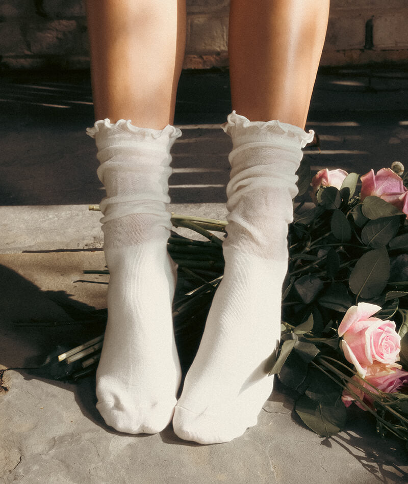 White socks with ruffles worn by a person, standing on a stone surface with flowers.