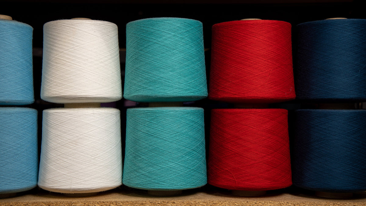 Eight large cones of thread or yarn in multiple colors, including light blue, white, teal, red, and dark blue, arranged in two rows on a wooden surface.