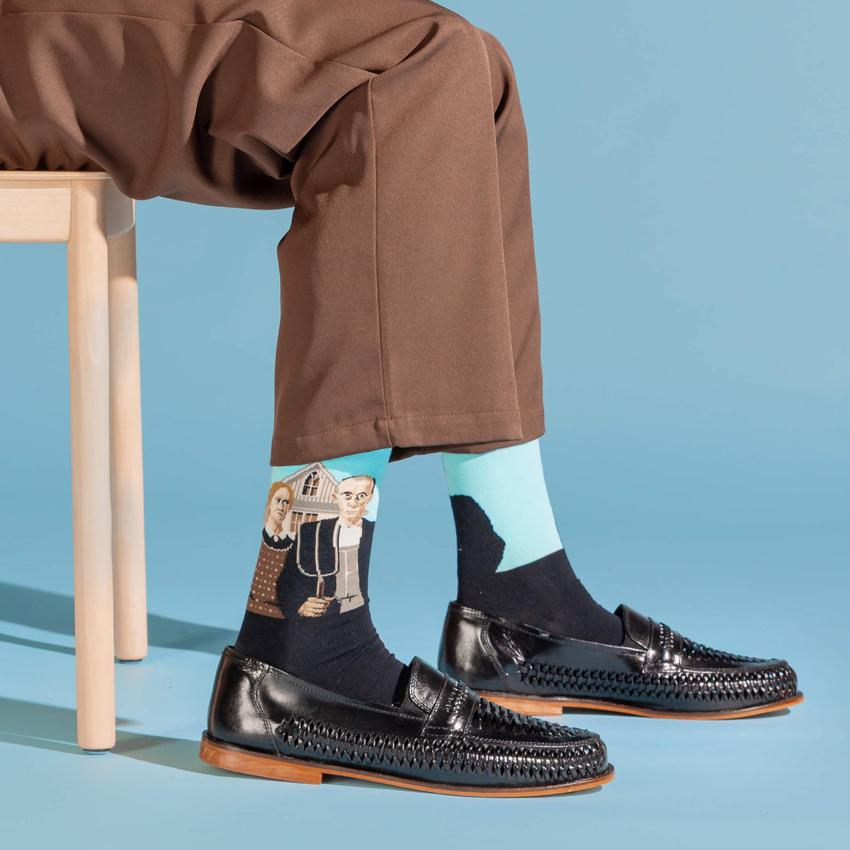 A person seated on a wooden stool wearing brown trousers, black woven loafers, and black socks with a colorful illustrated print. The background is a solid light blue studio setting.