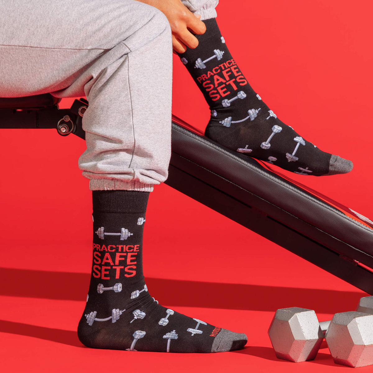 A person seated on a gym bench wearing black crew socks with red and white dumbbell graphics and the text 'PRACTICE SAFE SETS.' The scene is set against a bright red studio background with dumbbells on the floor.