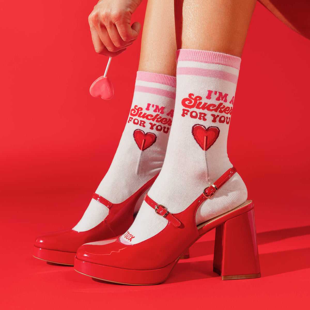White crew socks with red text reading 'I'M A Sucker FOR YOU' and a red heart, worn with red Mary Jane high heels on a bright red background. A hand holds a small pink heart-shaped object above the legs.
