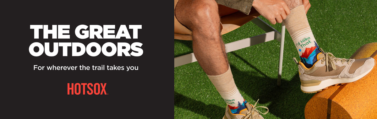 A promotional outdoor scene showing a person's legs and feet wearing beige hiking shoes and patterned socks on artificial grass, with large text on the left reading 'THE GREAT OUTDOORS' and 'HOTSOX'.