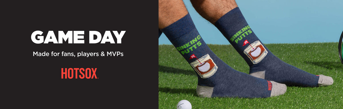 Dark navy blue athletic crew socks with a white and red HotSOX logo and green text, worn on a person's legs standing on a green grass field with a blue sky background. The left side of the image features a dark gray graphic with the text 'GAME DAY Made for fans, players & MVPs' and 'HOTSOX'.