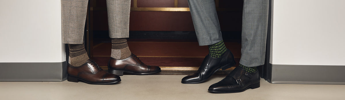A close-up of two people's lower legs and feet standing in a doorway, wearing brown and black leather dress shoes with different socks.