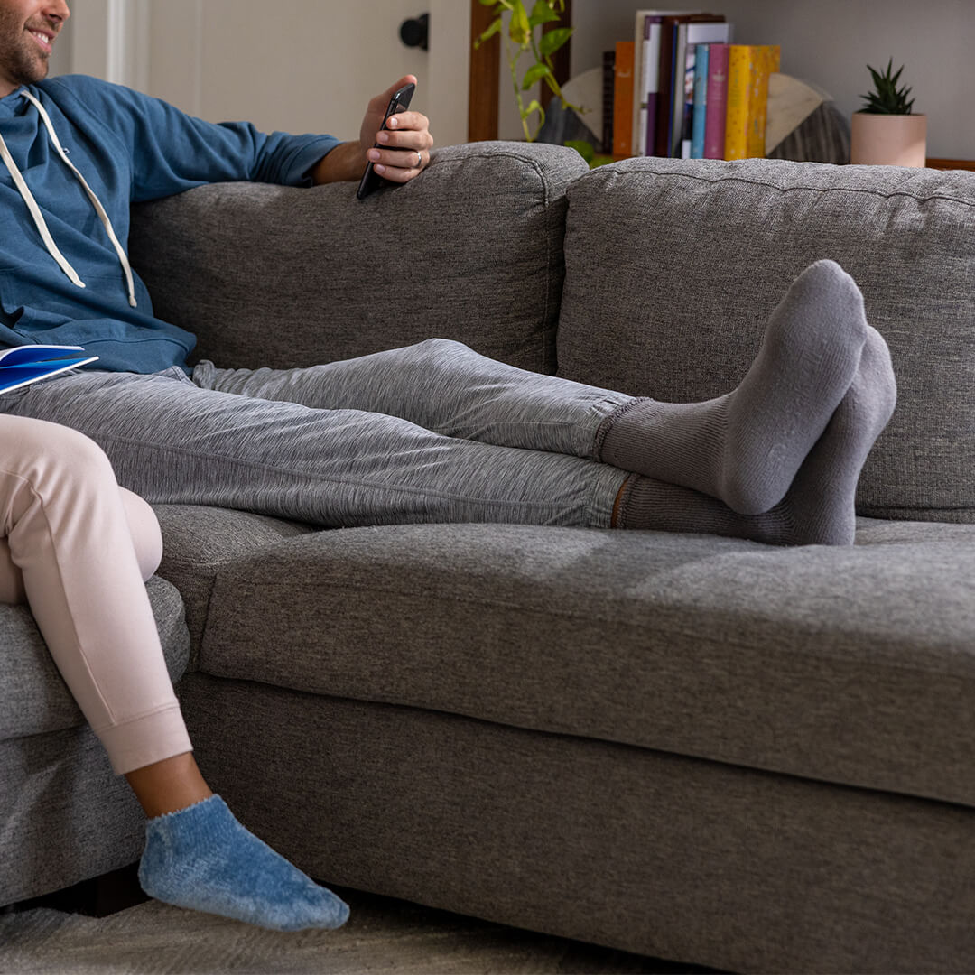 Person sitting on a gray couch wearing gray socks with another person wearing blue socks.