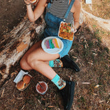 Person sitting outdoors with food, wearing denim overalls and colorful socks.